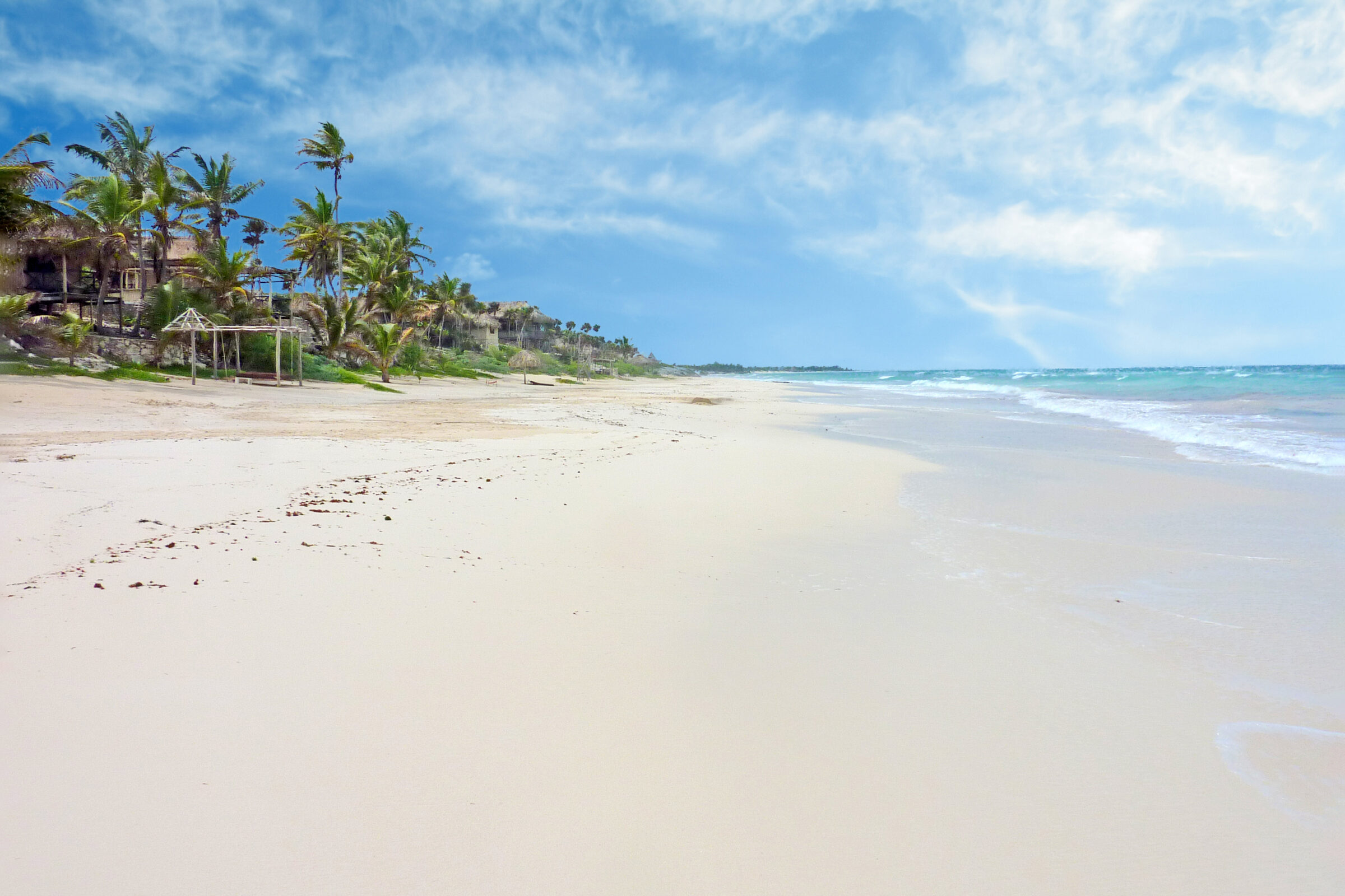 Plage de Tulum au Mexique avec sable blanc, mer turquoise des Caraïbes calme, palmiers luxuriants, ciel bleu ensoleillé et quelques nuages.
